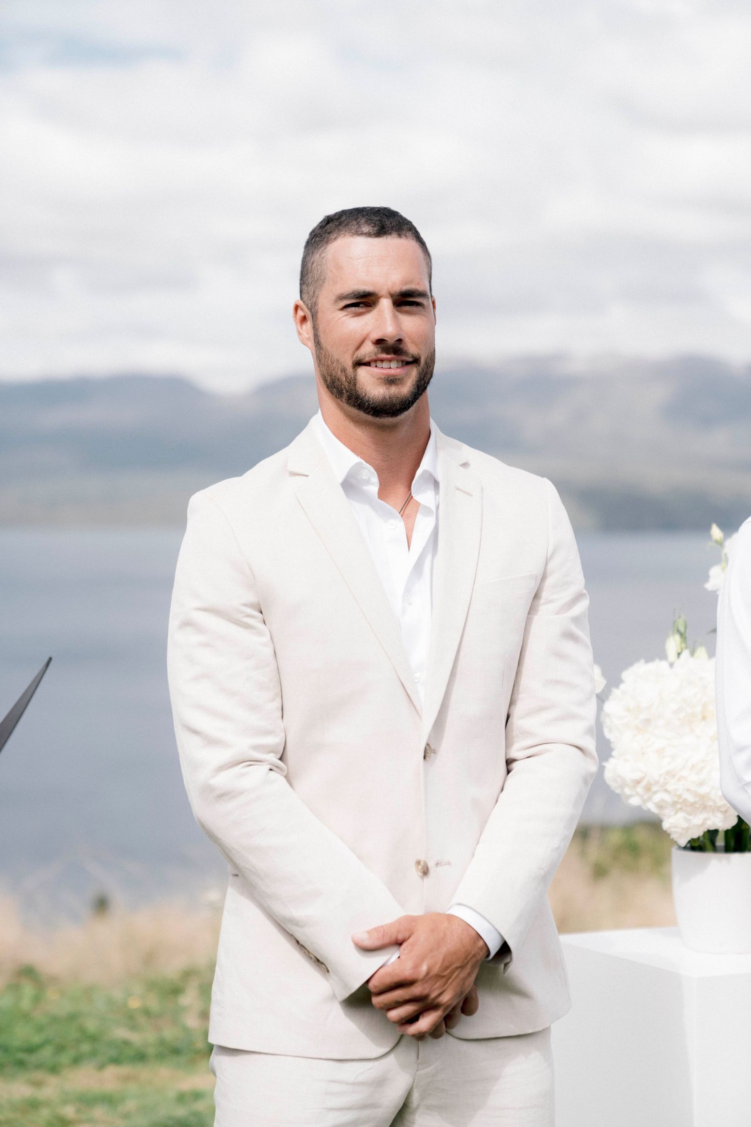 Groom in cream suit standing against mountain landscape with white floral arrangements.