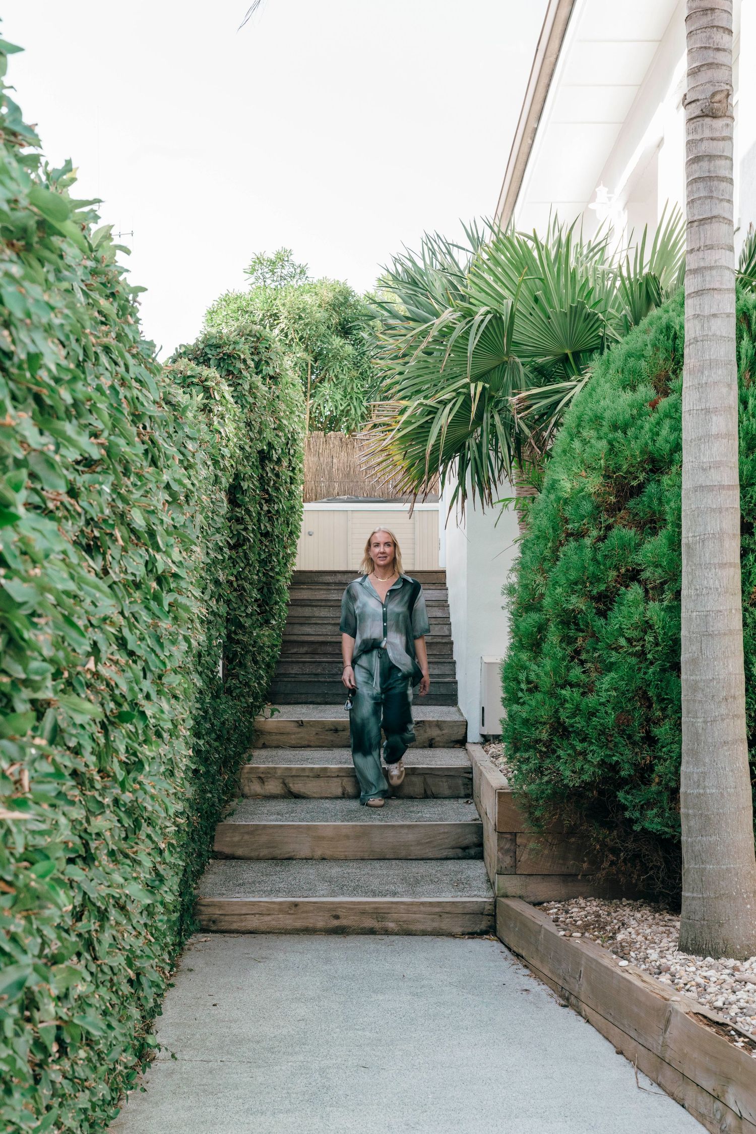 Concrete stairway flanked by lush green bushes and palm trees leading to a modern outdoor entrance.