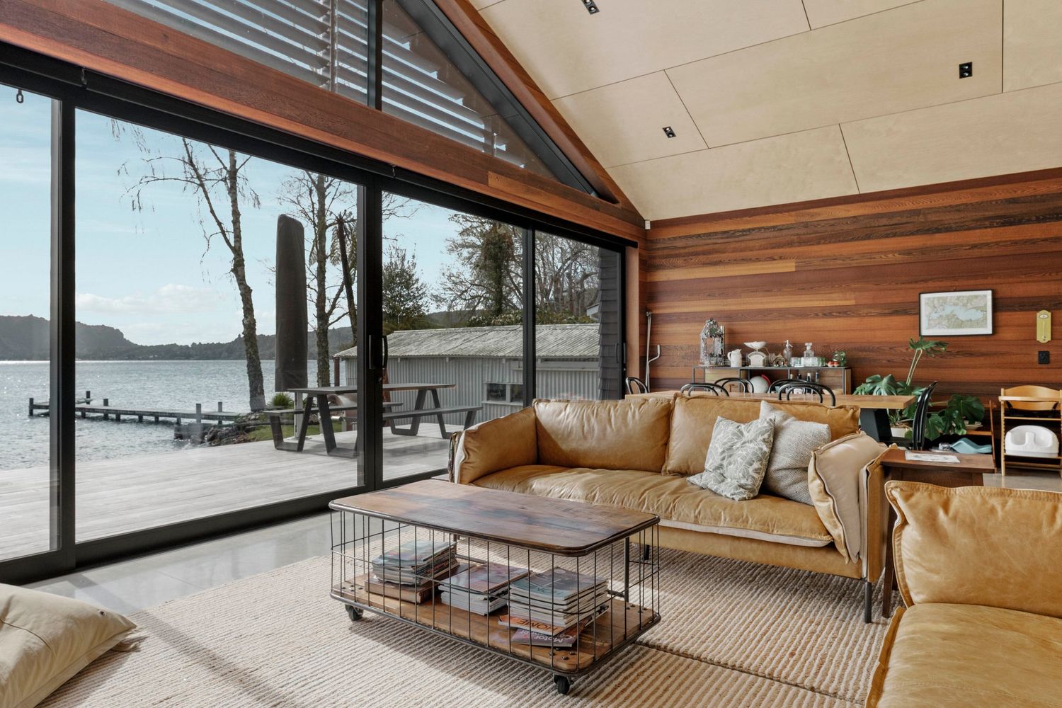 Modern lakefront living room with floor-to-ceiling windows, wood paneling, and tan leather furniture overlooking serene water views.