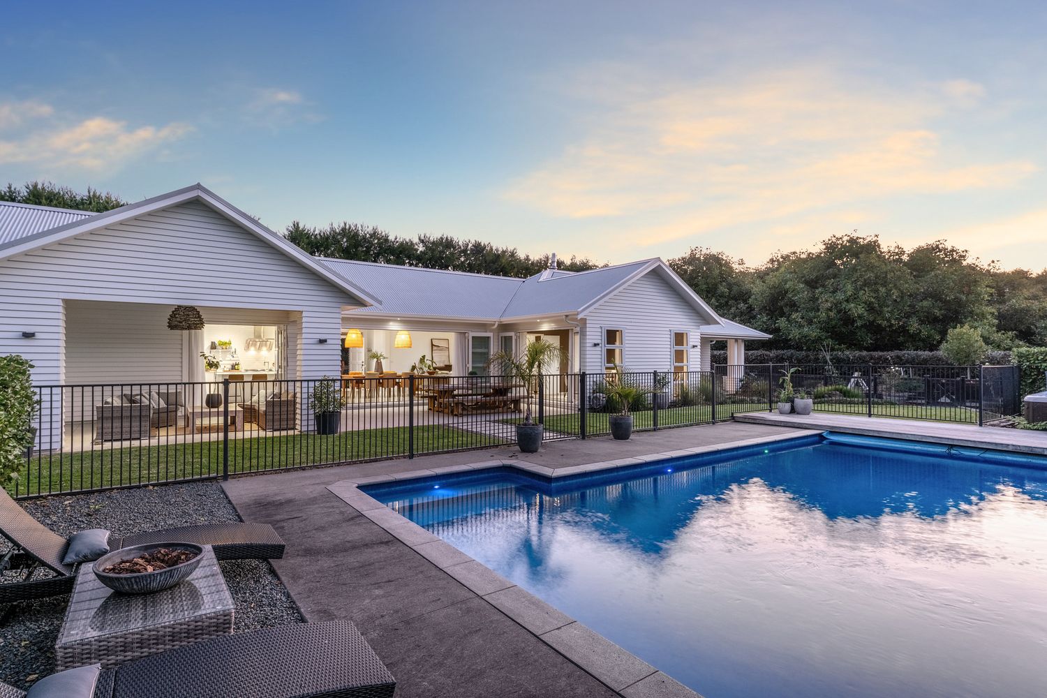 Modern white ranch-style home with fenced swimming pool and patio illuminated at dusk.