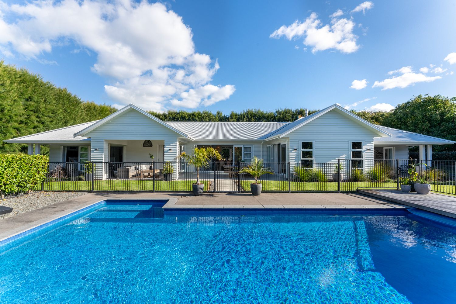 Panoramic view of a pristine blue swimming pool next to a white residential building under blue skies.