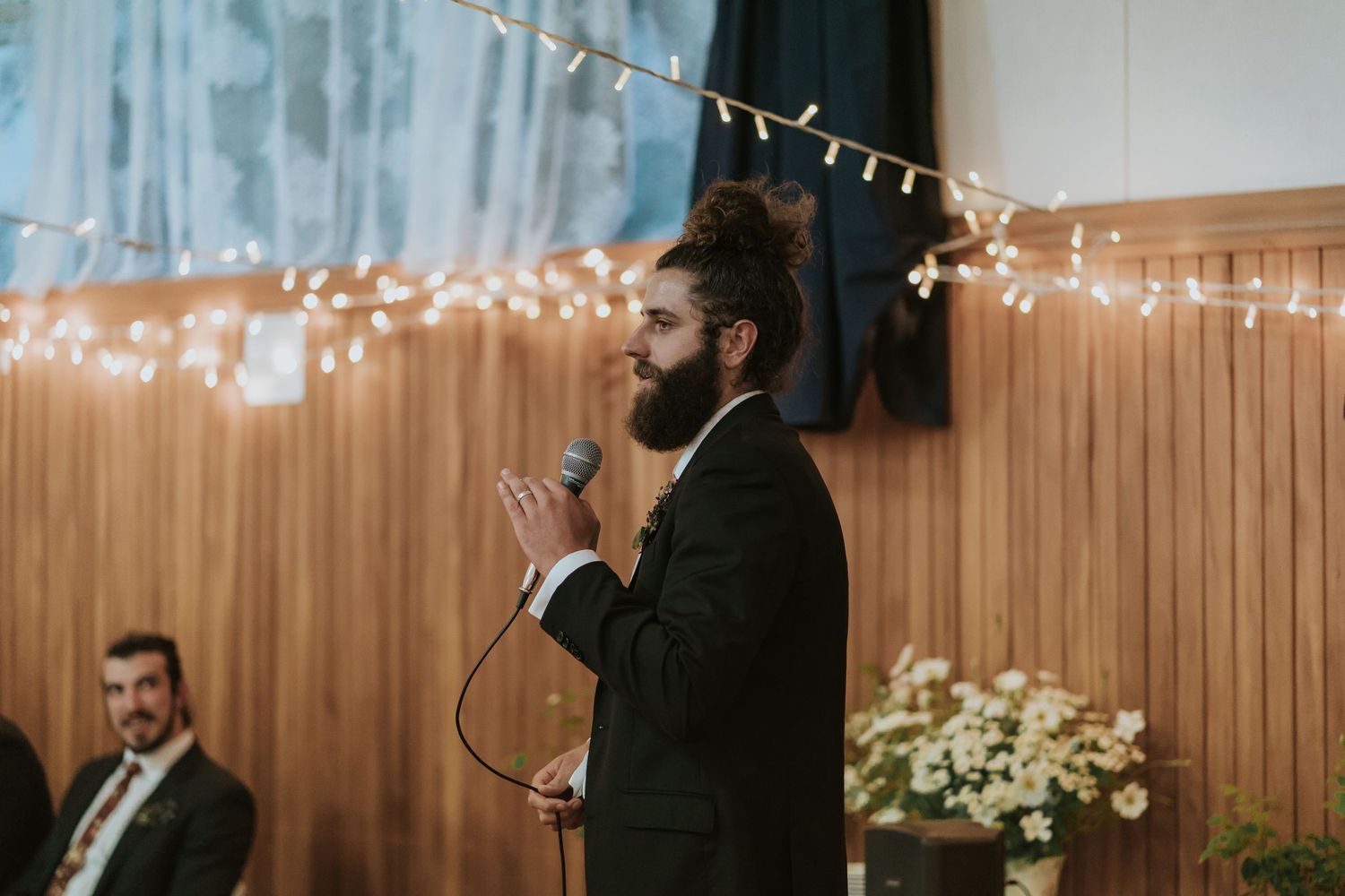 A person speaks into a microphone at an indoor event space decorated with string lights and wooden paneling.