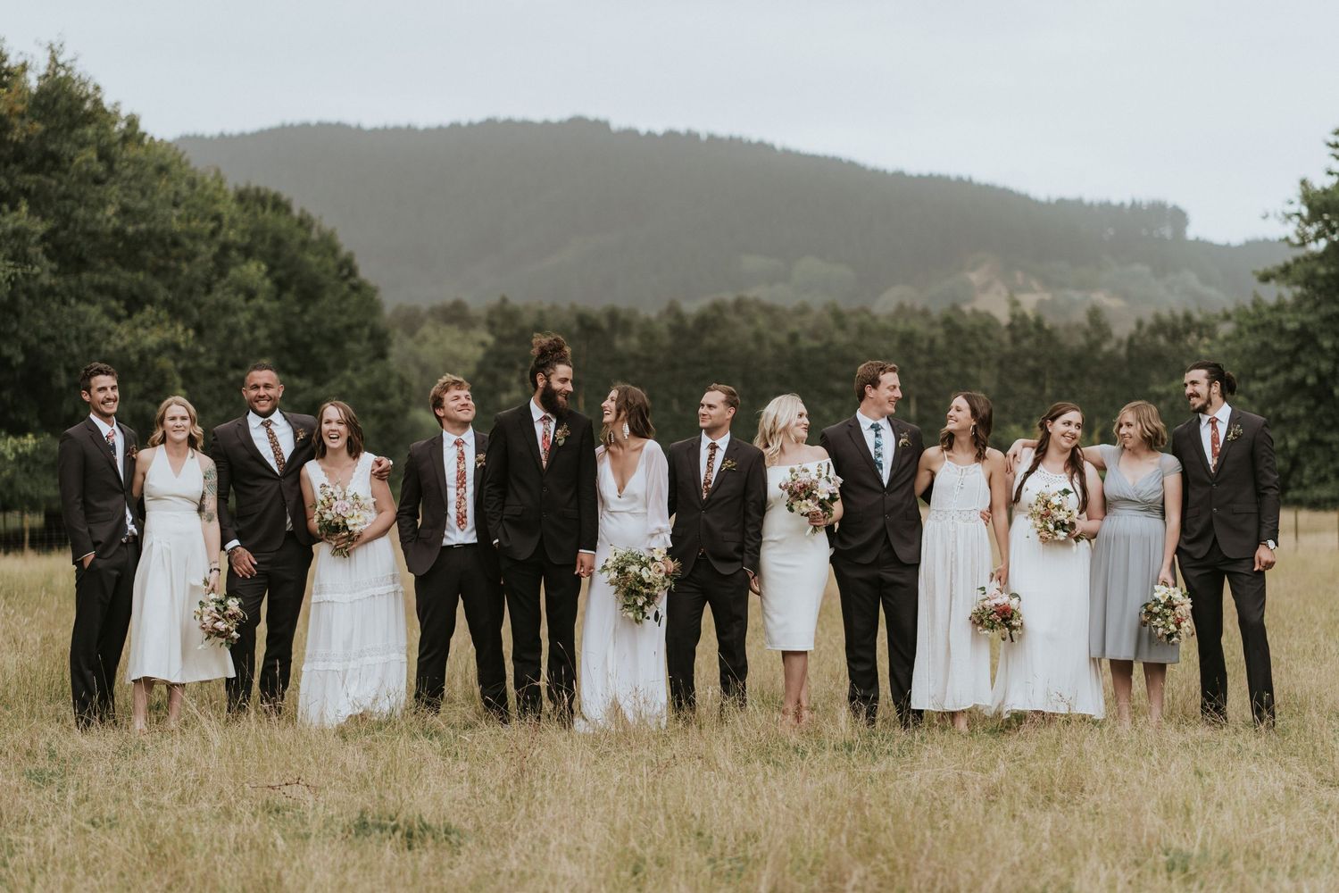 Wedding party in coordinated white and black attire poses together in golden field with mountains in background.