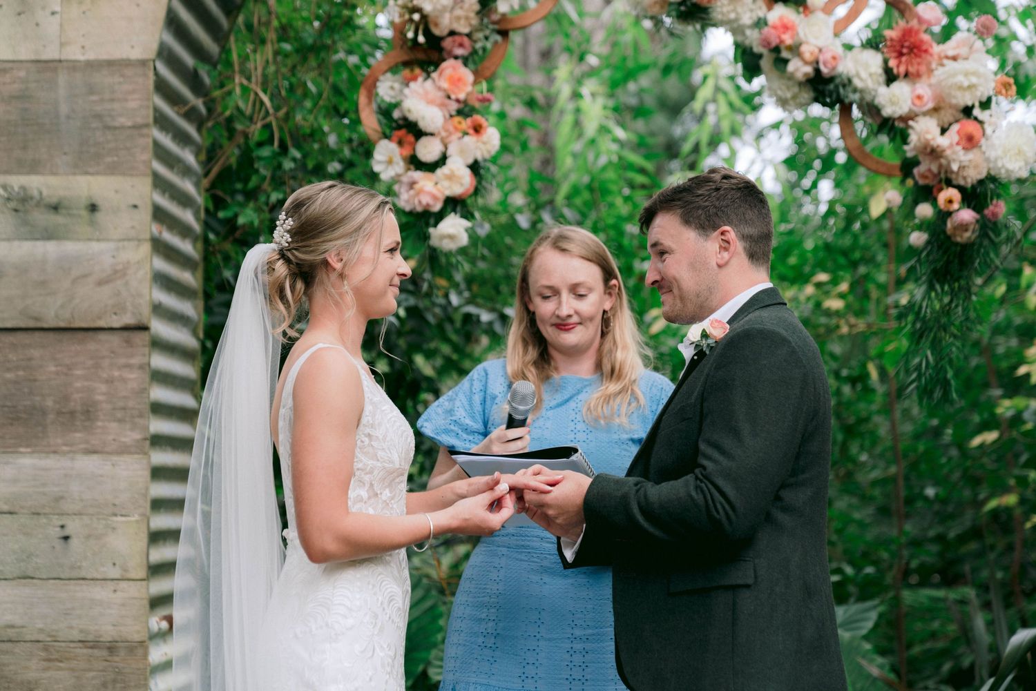 Outdoor wedding ceremony under trees with floral decorations and couple exchanging rings.