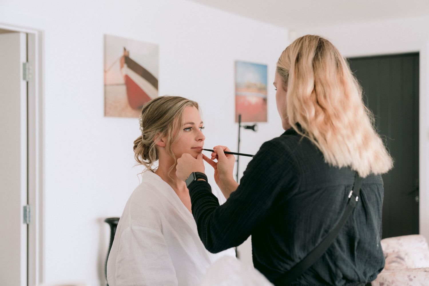 Professional makeup artist in black attire applies cosmetics on client in bright studio setting.