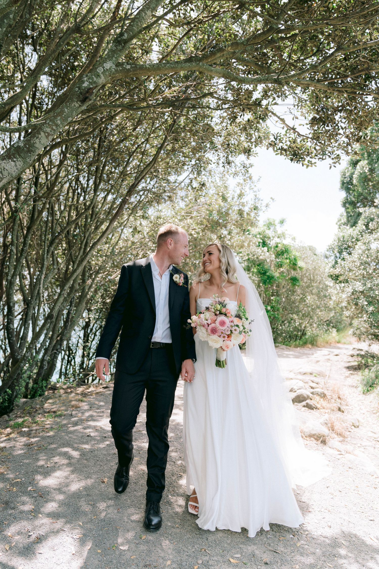 A couple walking together on a tree-lined path during their wedding day, with blooming trees creating a natural canopy.