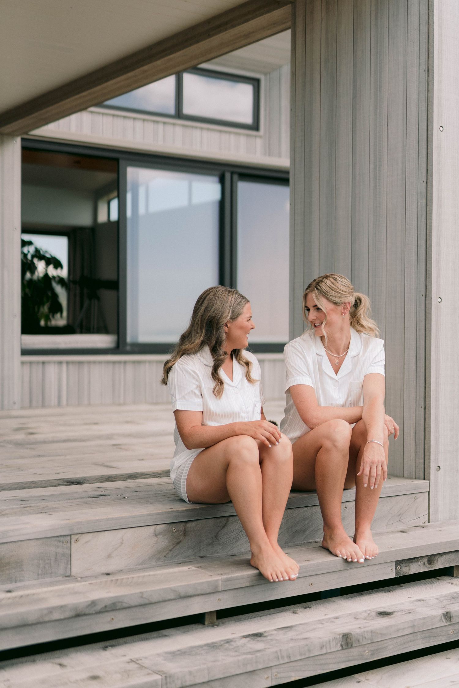 Two people in white outfits sit casually on wooden deck steps while having an intimate conversation outdoors.