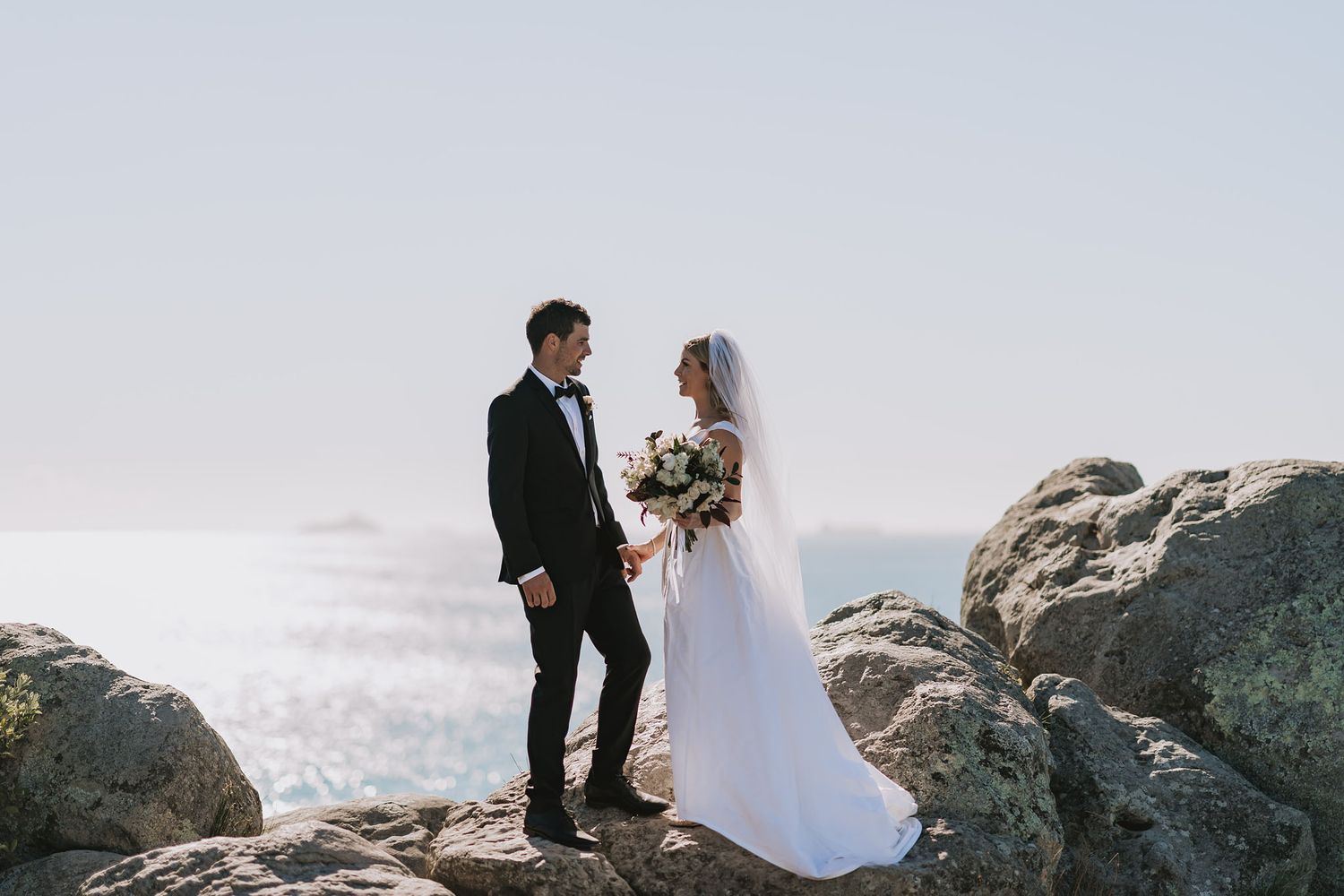 Scenic coastal wedding portrait with couple standing together on rocky cliffs by the ocean.