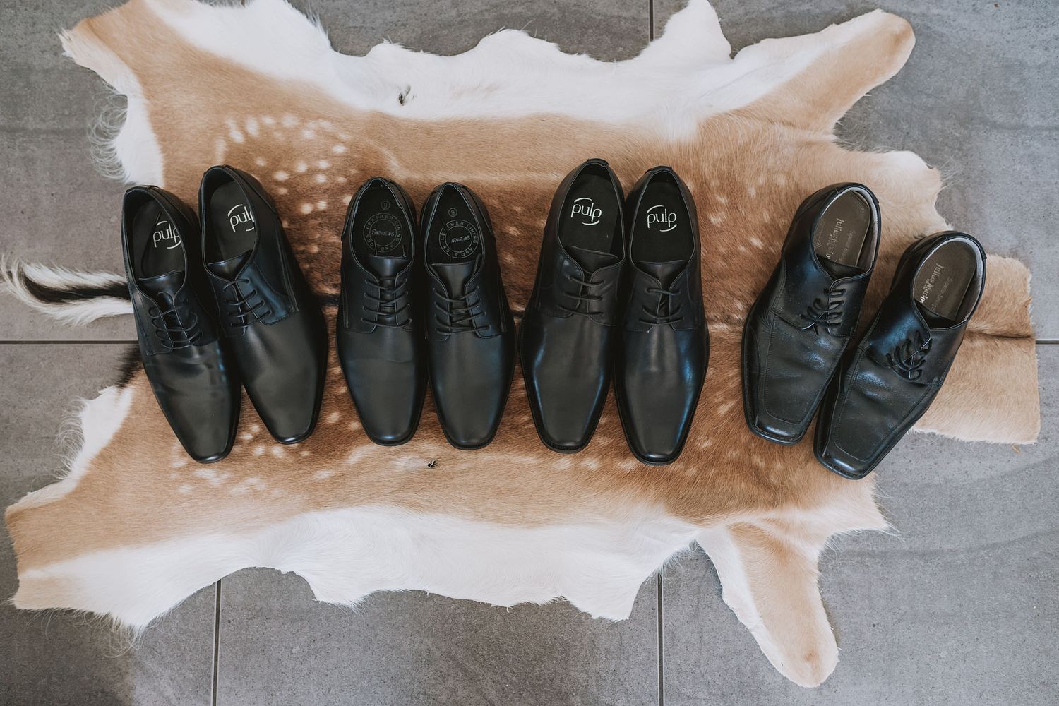 Four pairs of black leather dress shoes arranged in a row on a beige cowhide rug against a grey floor.