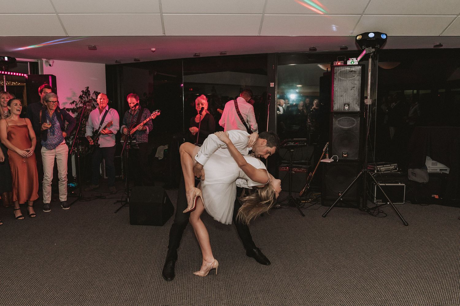 A couple performs an energetic dance move at a party venue with pink lighting and a band in the background.