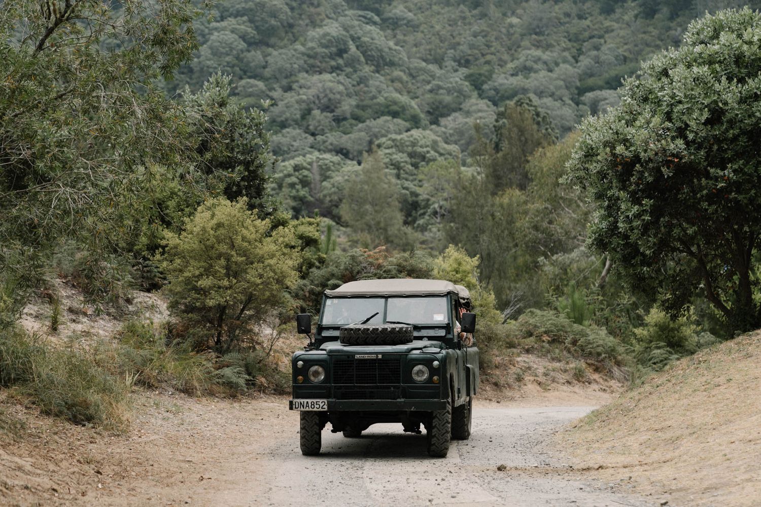 Black vintage Land Rover Defender driving on dirt road through green mountainous terrain.