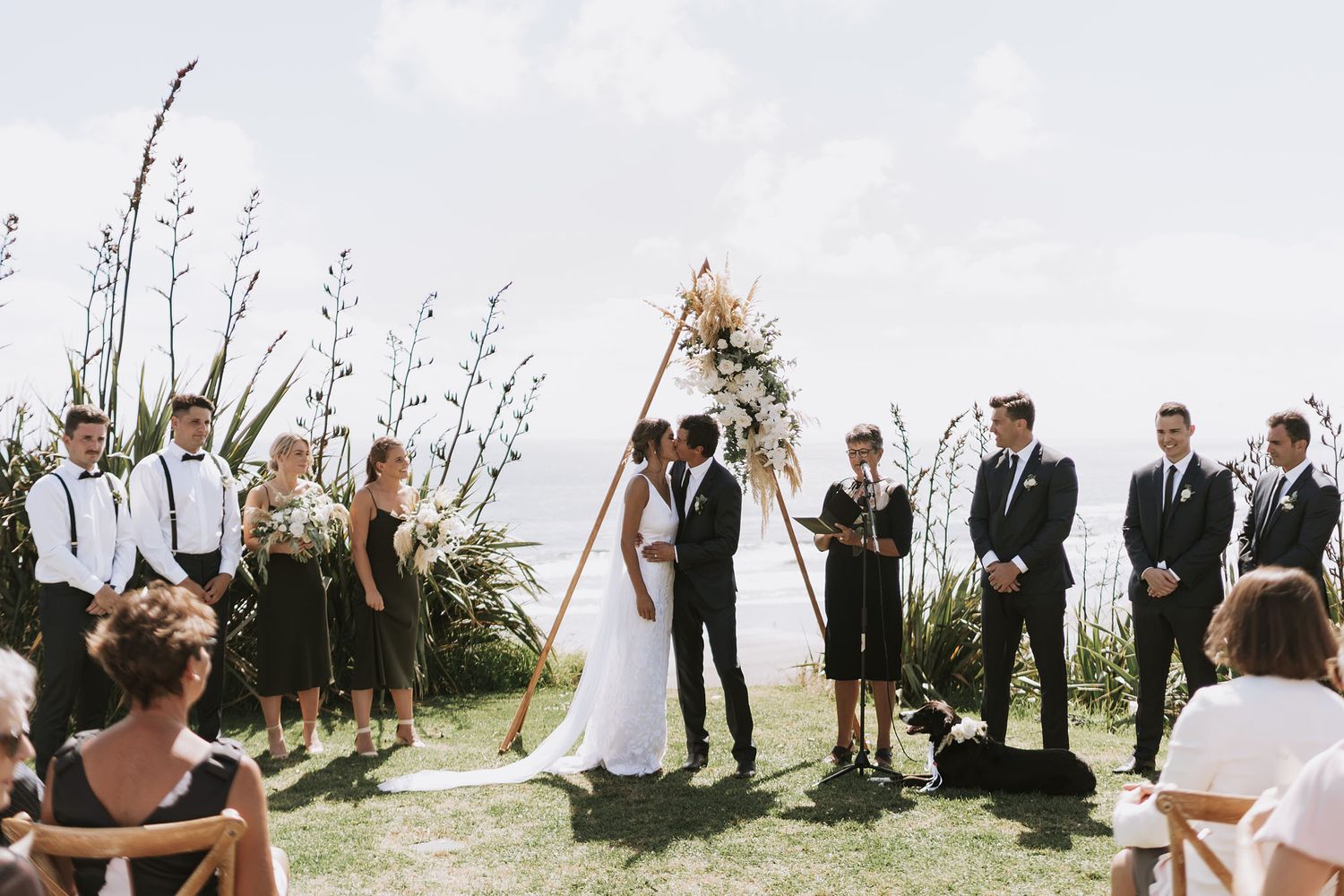 Wedding party stands in a line against bright sky during outdoor ceremony with groomsmen and bridesmaids.
