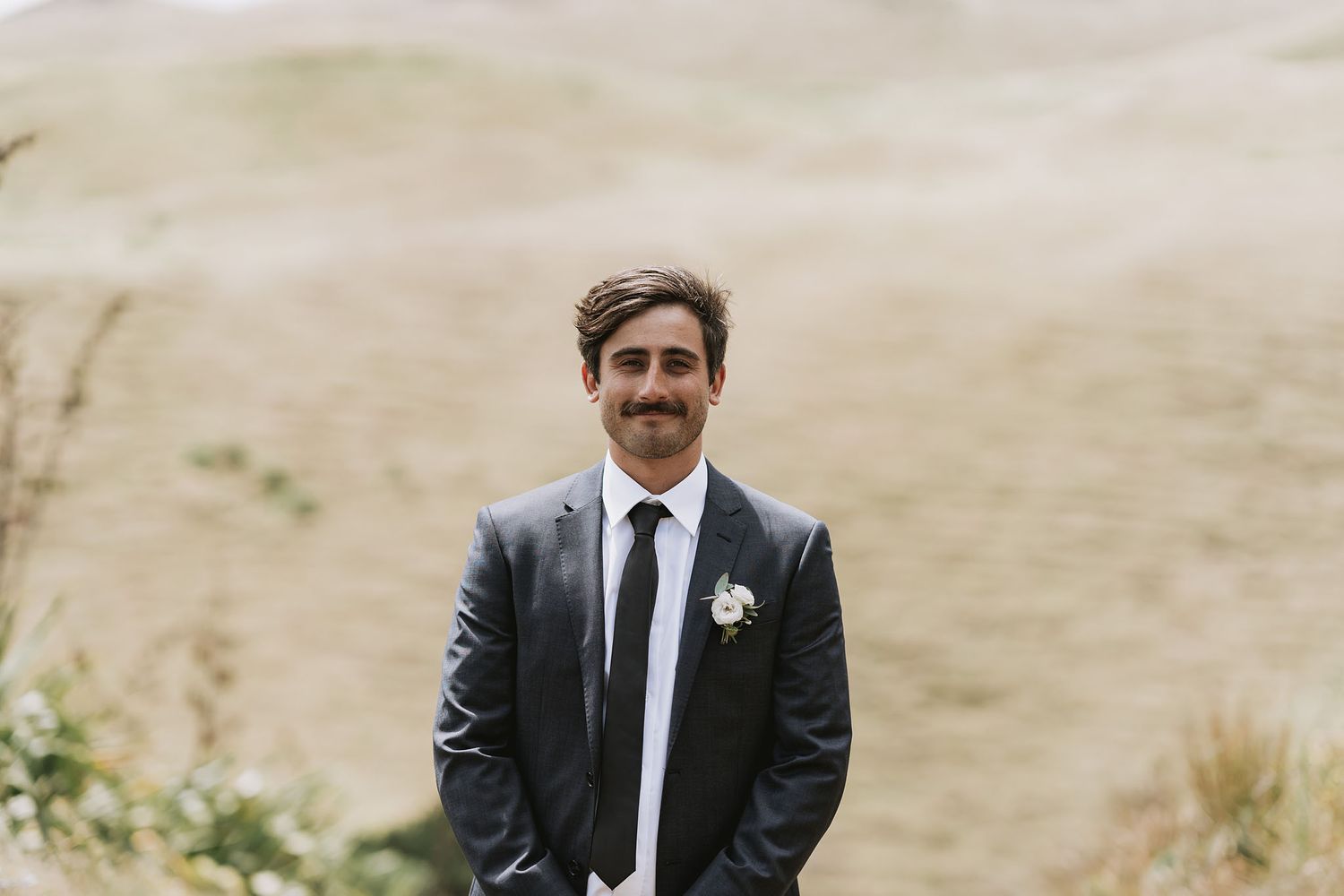 A group of six groomsmen in black suits and ties pose together in a rustic outdoor setting with golden grass.