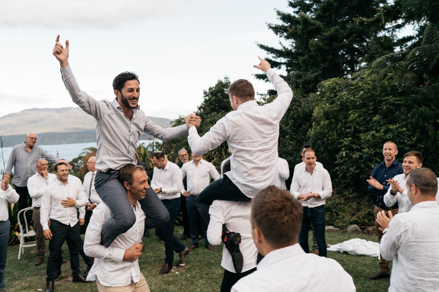 Two people jump excitedly while surrounded by guests during an outdoor celebration with mountains in the background.