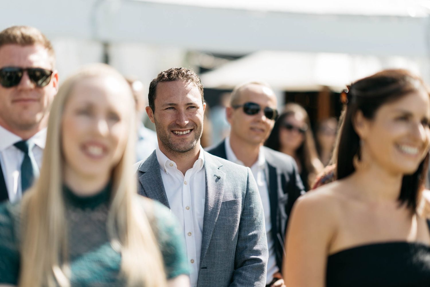 A cheerful outdoor wedding gathering with guests in formal attire smiling and mingling during a sunny reception.