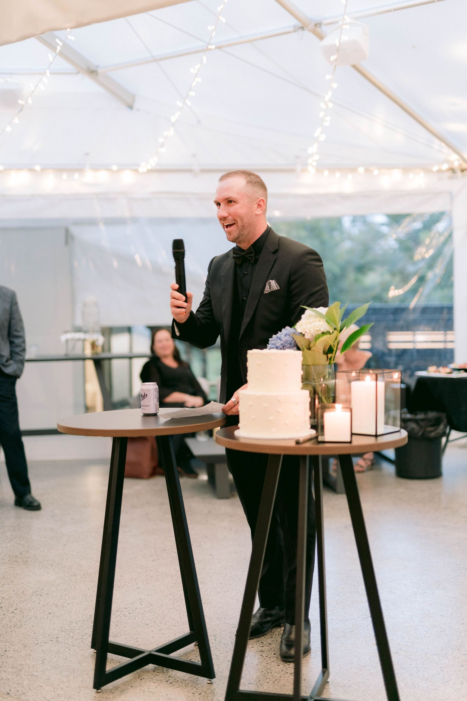 Person in black suit seated at table with cake under white tent with string lights.
