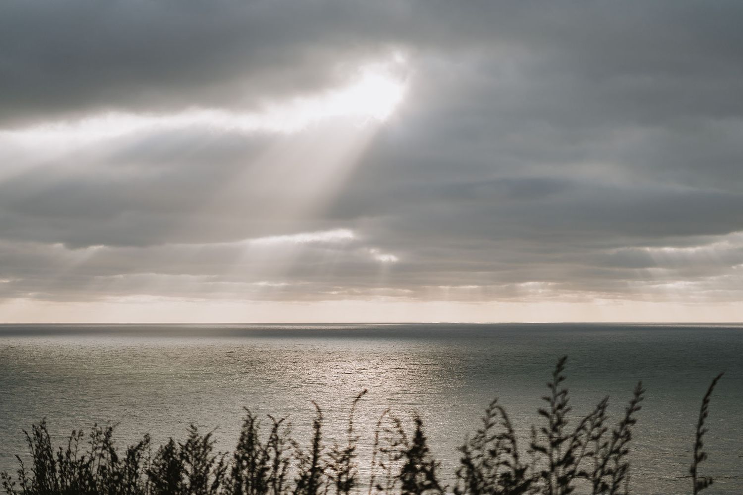 Dramatic stormy skies cast sunbeams over a dark and moody ocean landscape at sunset with dramatic cloud formations.