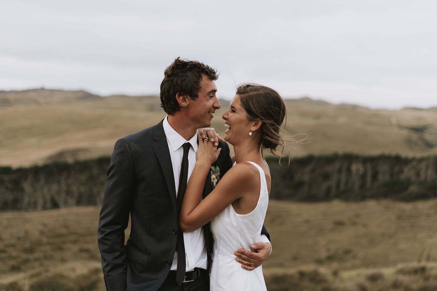 A couple in wedding attire embraces against a scenic mountainous landscape at sunset.