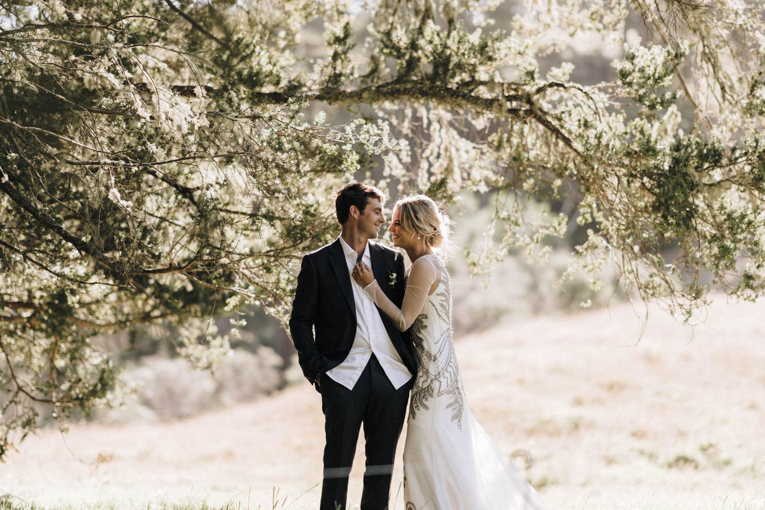 Romantic couple in wedding attire embraces under sunlit tree branches creating a dreamy outdoor atmosphere.