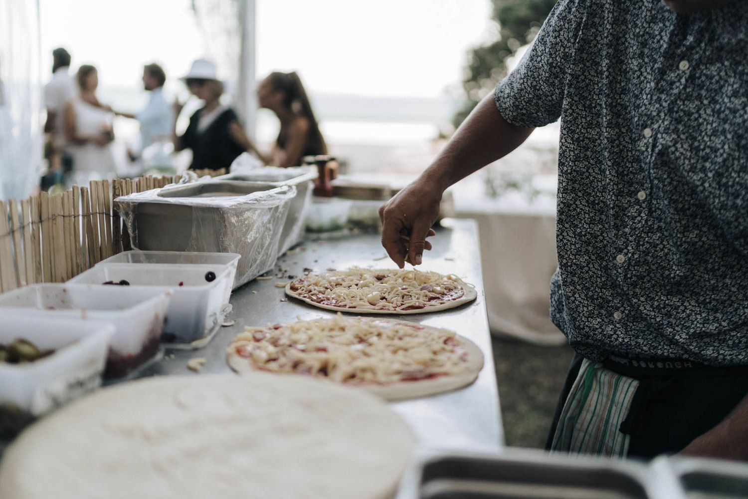 Chef preparing multiple pizzas at a rustic outdoor catering event with wooden tables in the background.