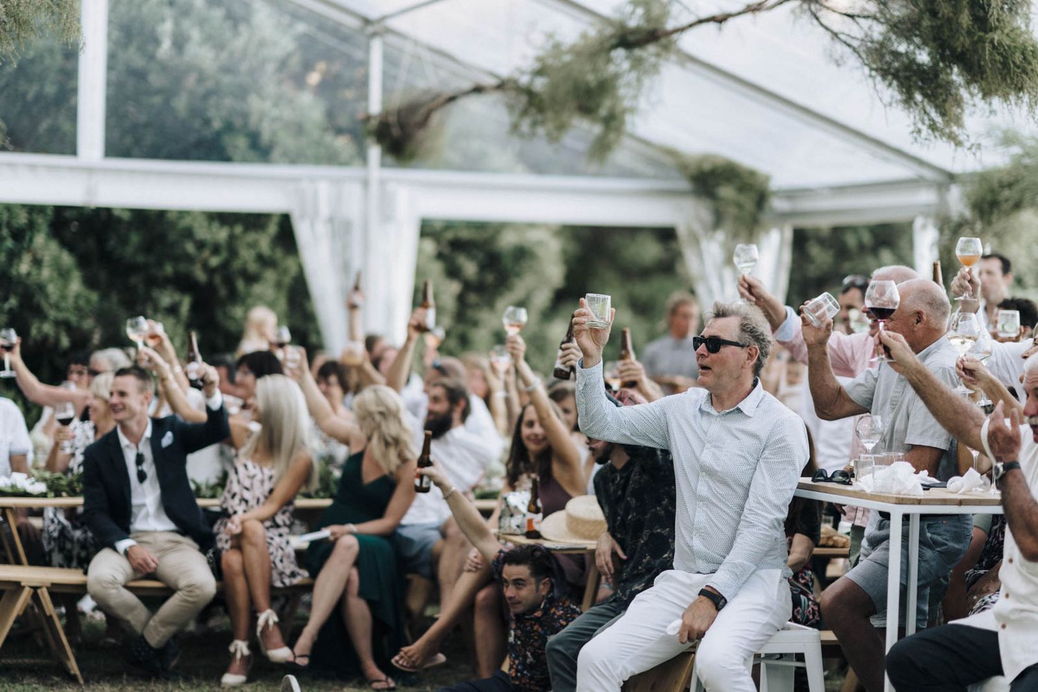 Wedding guests raising their hands and dancing joyfully during an outdoor celebration under a white tent.