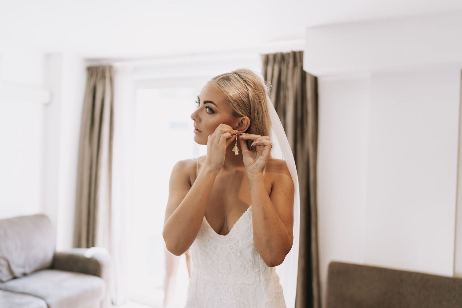 A bride in a white wedding dress puts on earrings while getting ready in a light-filled room with a gray sofa.