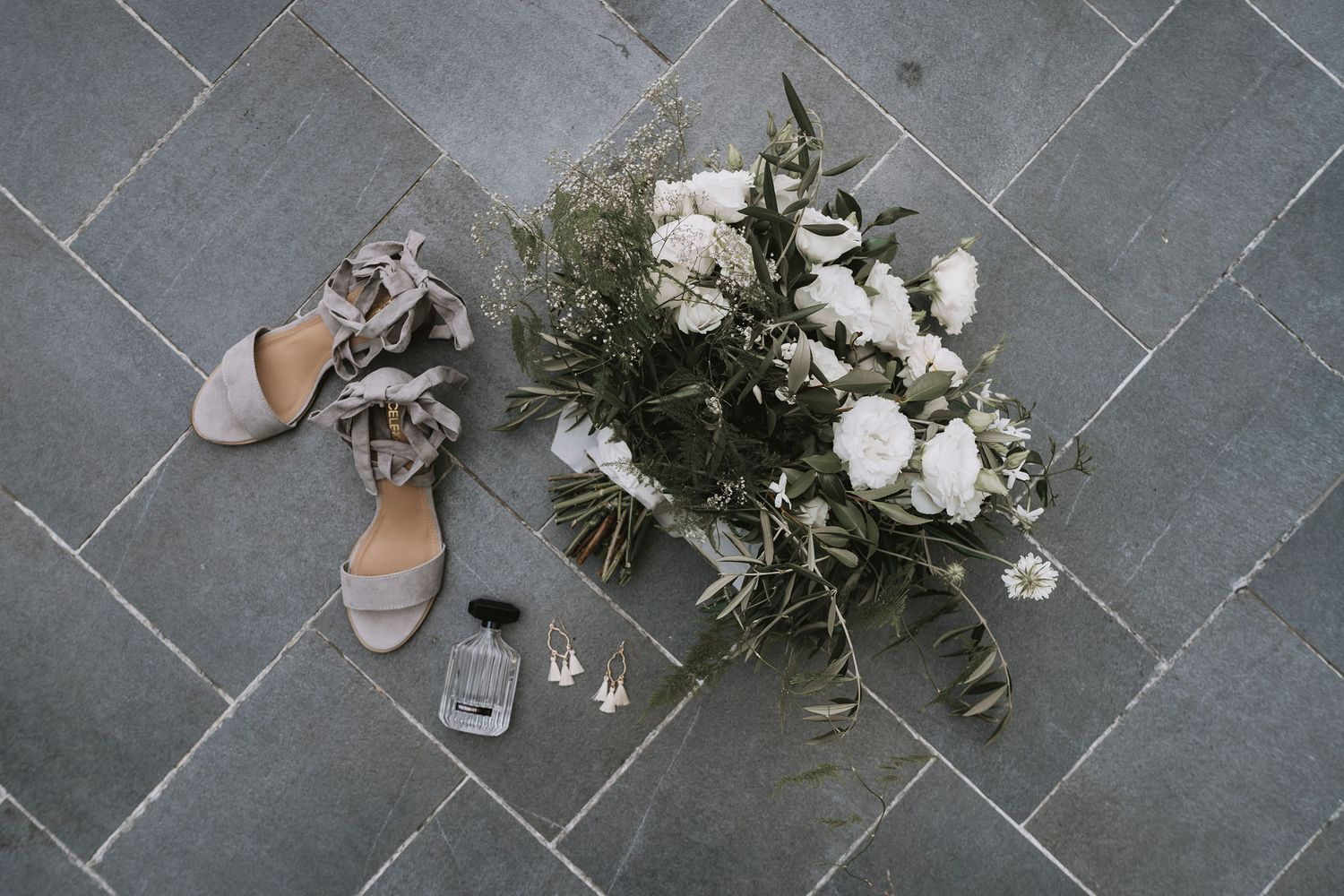 Wedding accessories including elegant sandals, delicate floral bouquet and glassware arranged on gray tile flooring.
