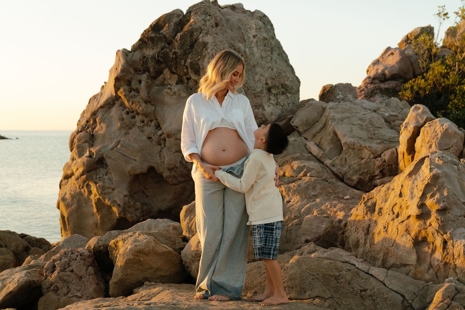 Pregnant woman in white shirt on rocky beach with child touching her belly at golden sunset. Bryony Alexander Newfound.