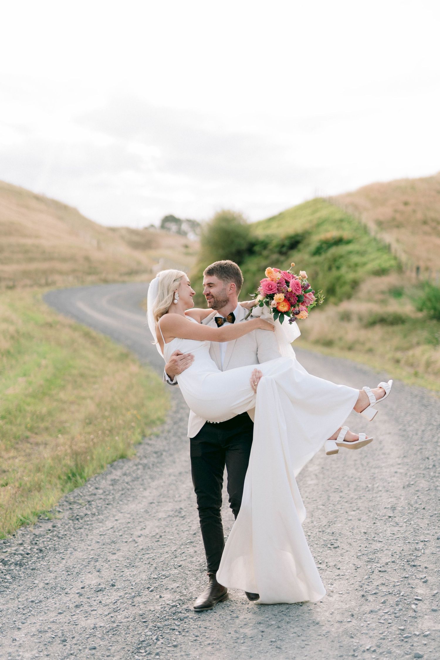 Newlyweds dance and twirl playfully on a winding dirt road surrounded by rolling green hills.