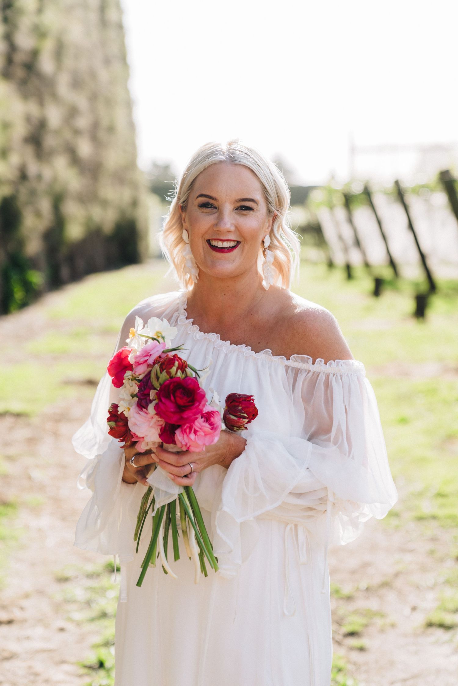 Wedding bouquet with vibrant pink and red flowers held against flowing white dress outdoors.