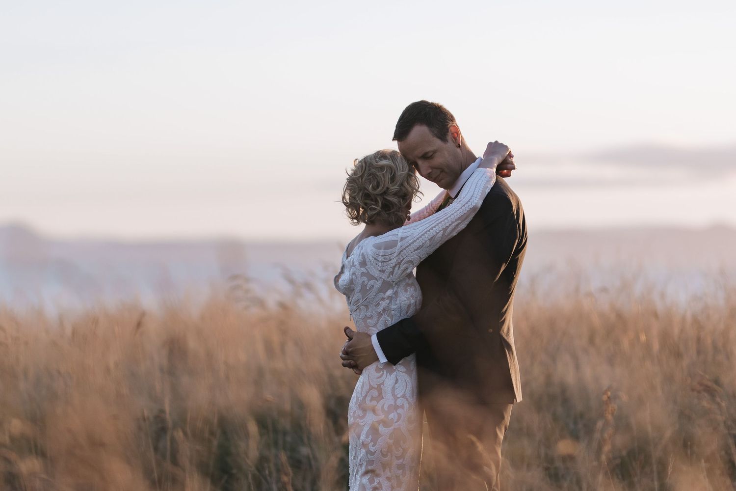 A couple in wedding attire share an intimate dance at sunset in a field of tall grass during golden hour.