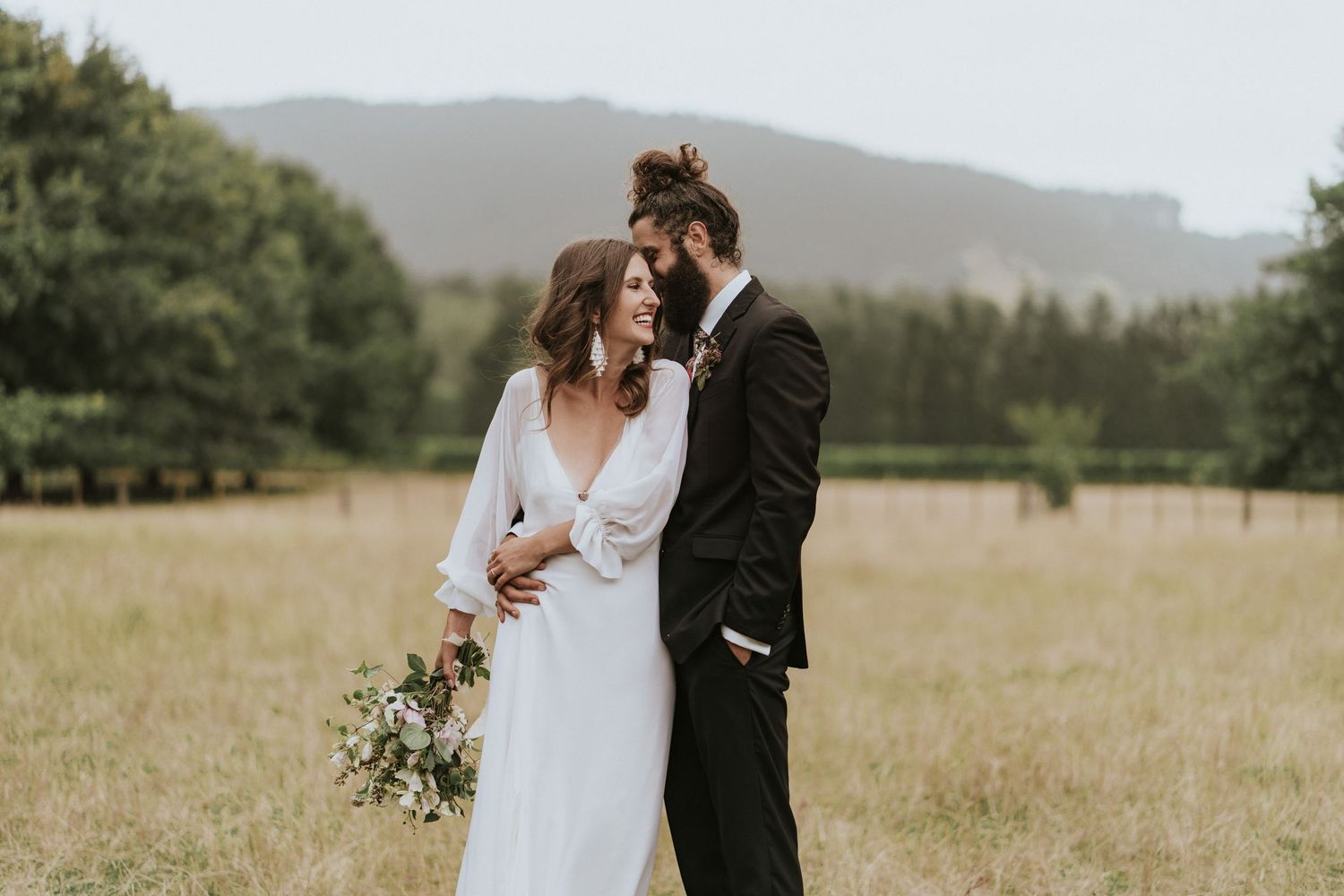 Couple shares intimate moment in elegant wedding attire against scenic mountain backdrop and grassy field.