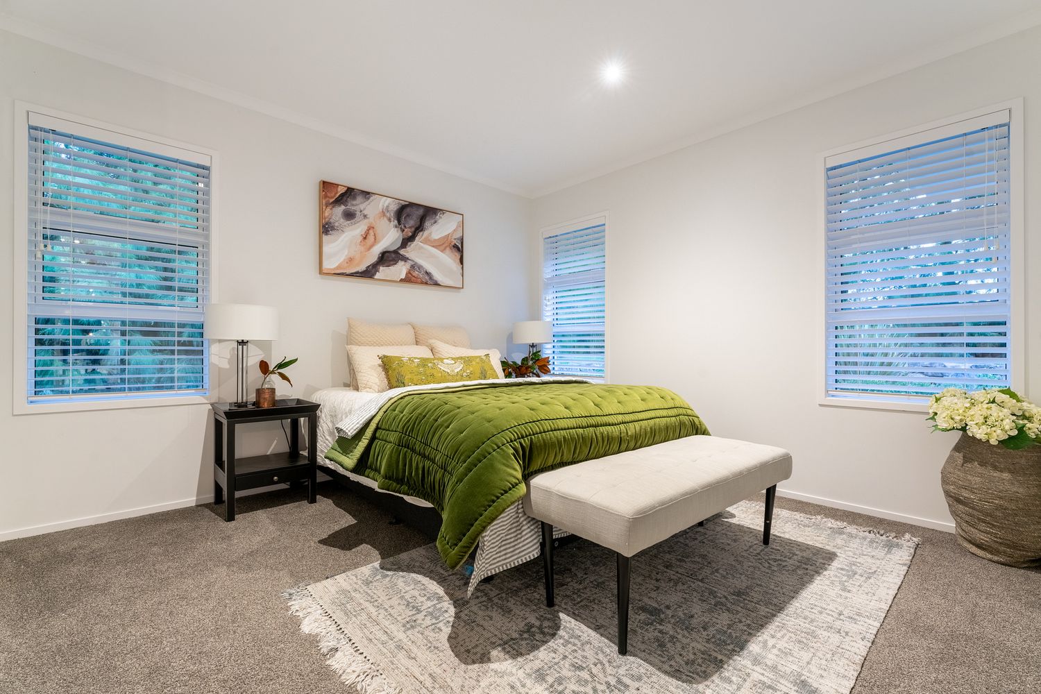 Contemporary master bedroom with grey carpet, green bedding, and dual windows featuring natural light.