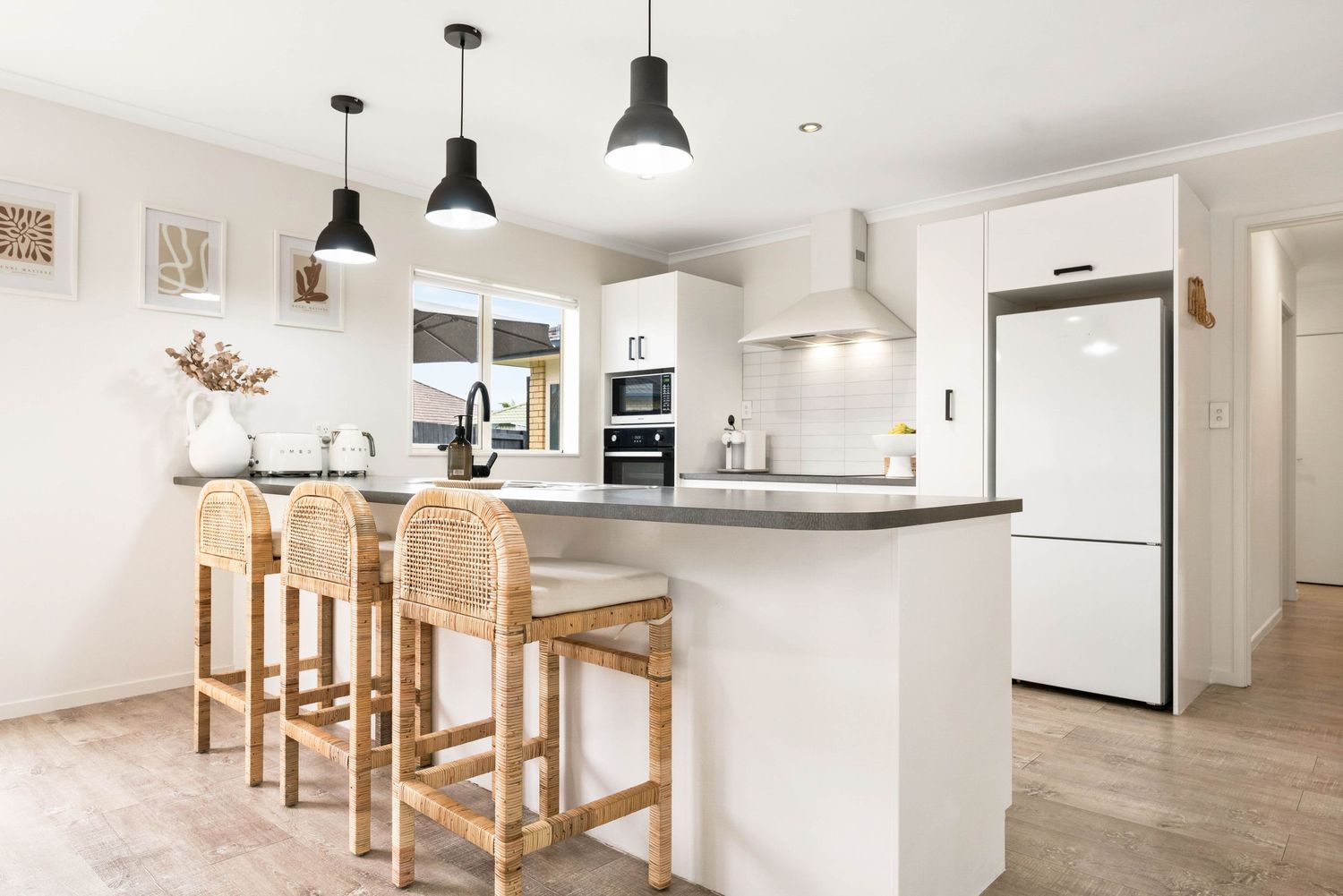 Modern white kitchen with pendant lights, wood bar stools, and grey countertops features a minimalist design.