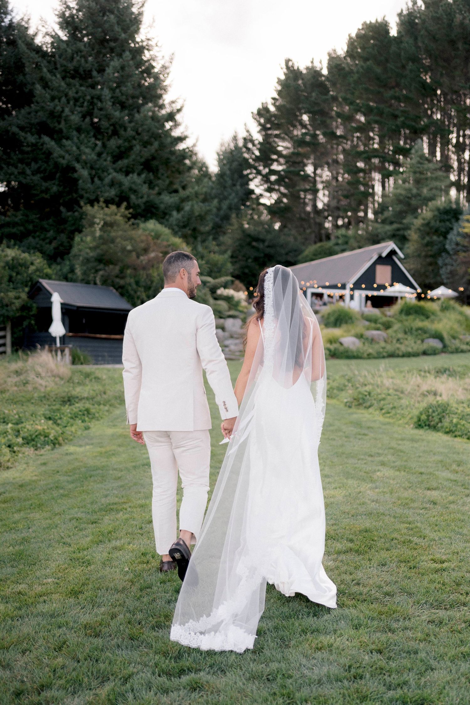 Bride and groom walking hand in hand on grass with evergreen trees in background.