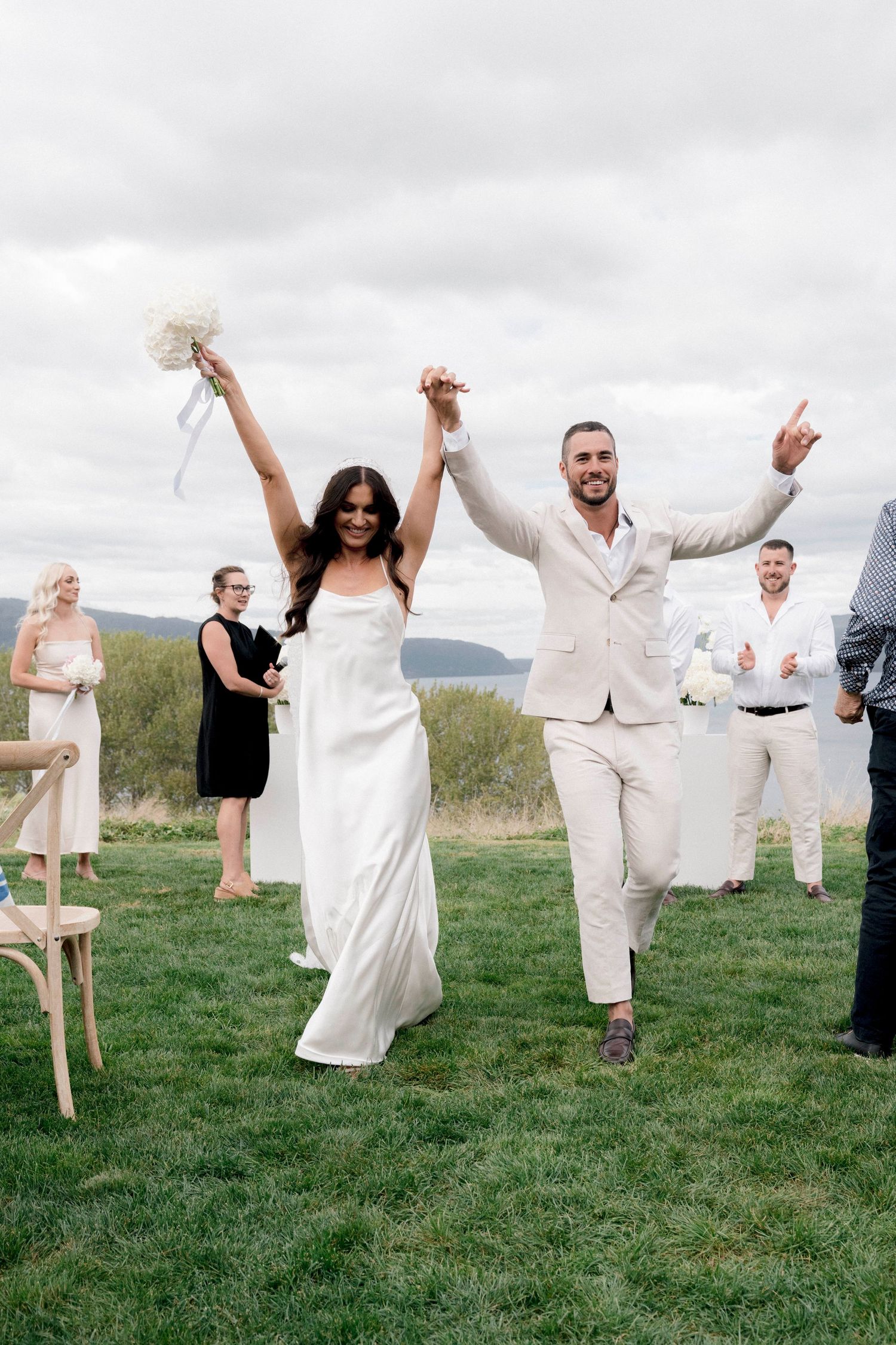 Jubilant newlyweds celebrate with raised arms after outdoor wedding ceremony on grassy hillside.