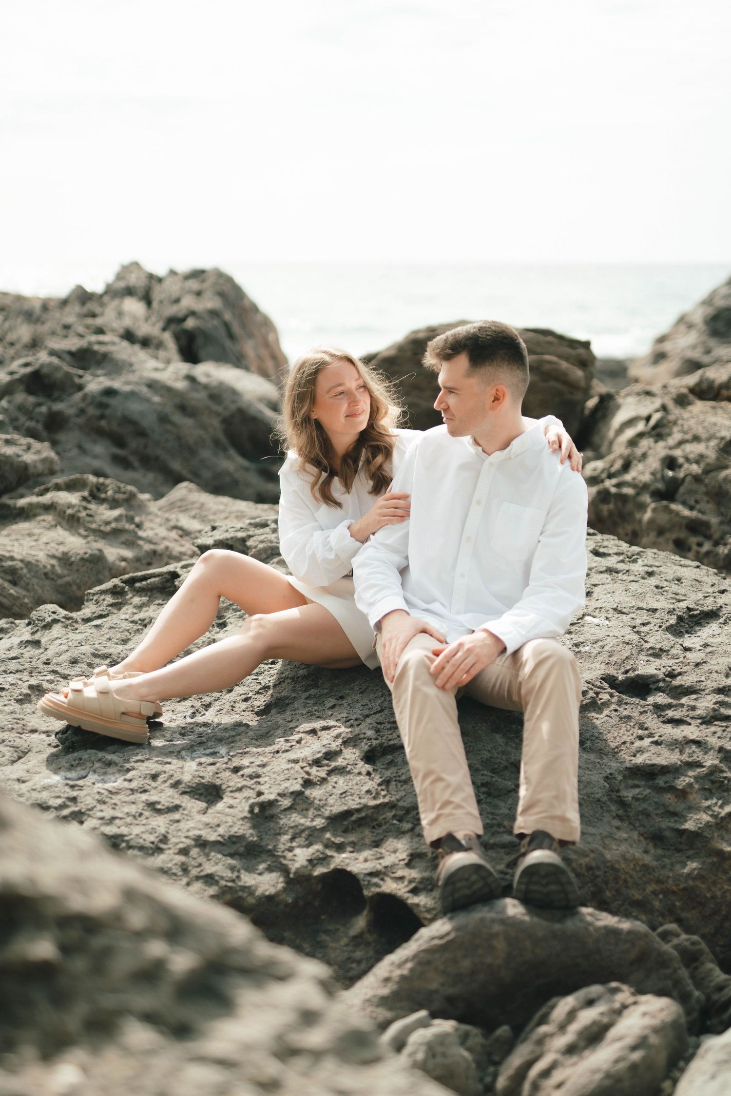 Intimate moment between couple relaxing on coastal rocks wearing matching white outfits.