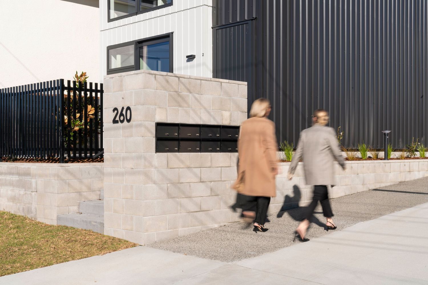 Two people in coats walk past a modern building entrance with address number 260 and black metal siding.