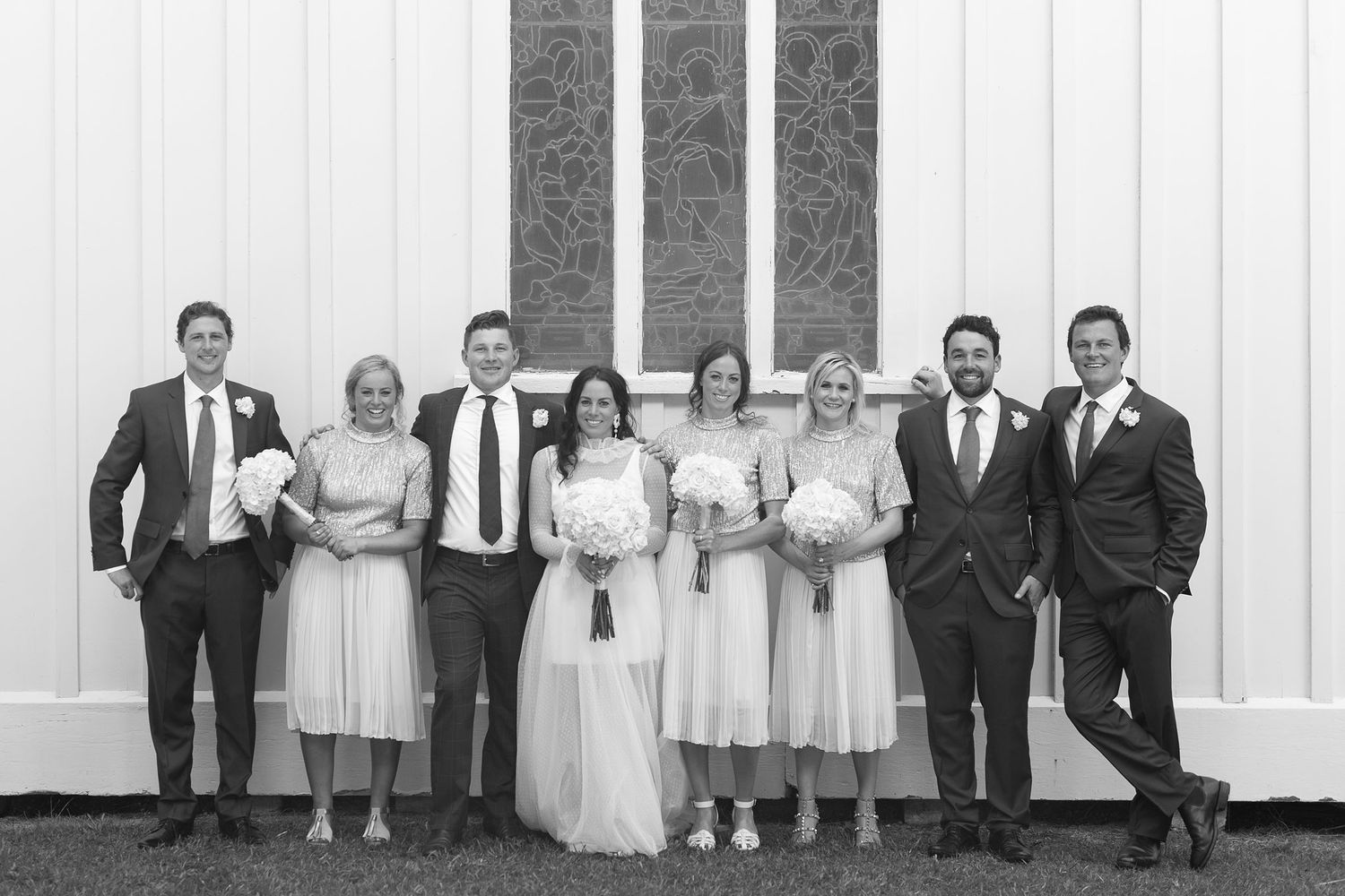 A wedding party lines up against a white wall wearing formal attire in black and white photo.