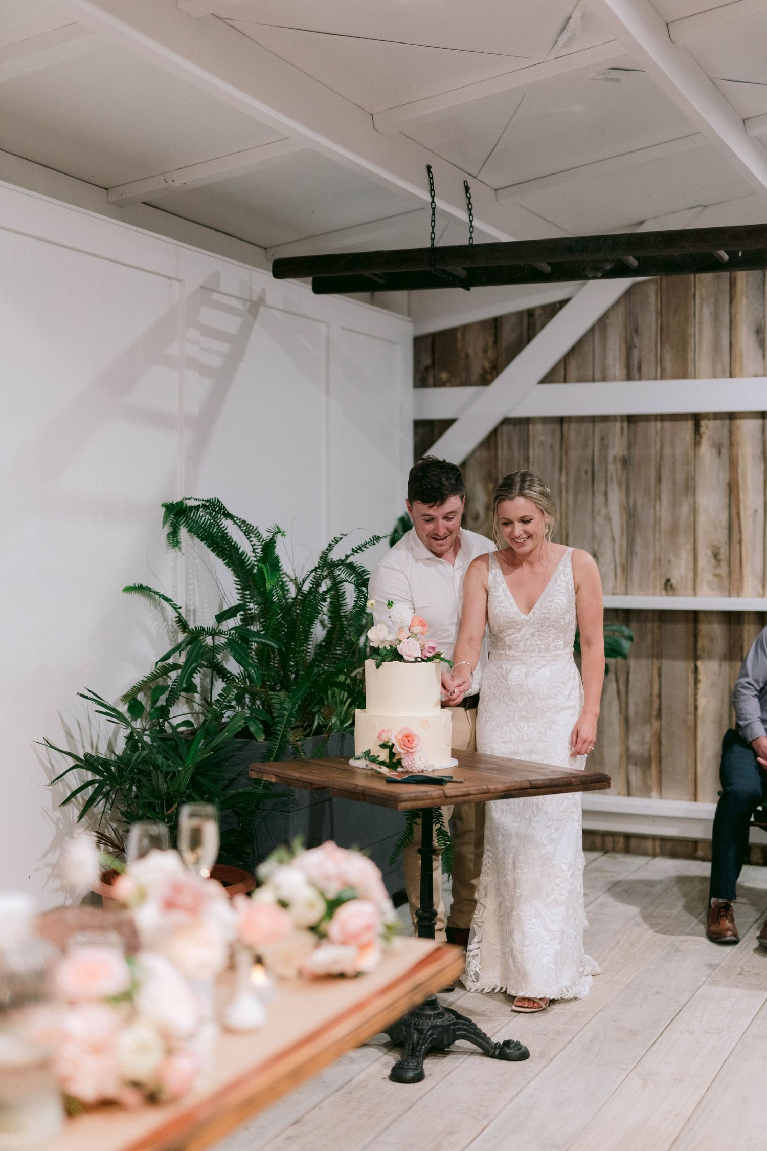 A wedding couple stands together near their elegant white cake decorated with greenery and flowers.
