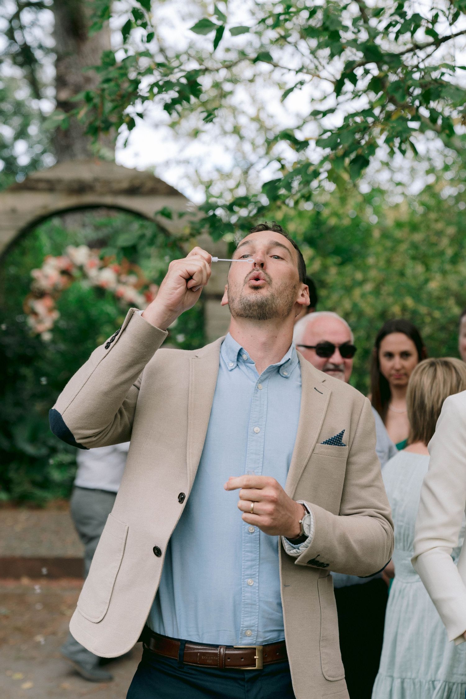 Wedding guest in beige blazer blowing bubbles during outdoor celebration.
