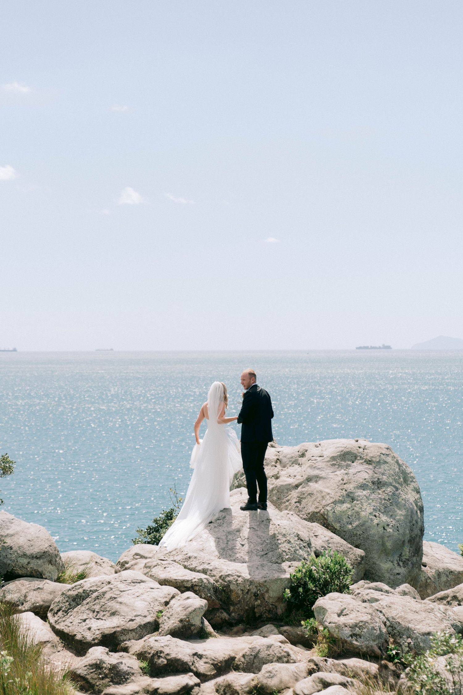 Wedding couple standing on rocky coastal outcrop with ocean view backdrop.
