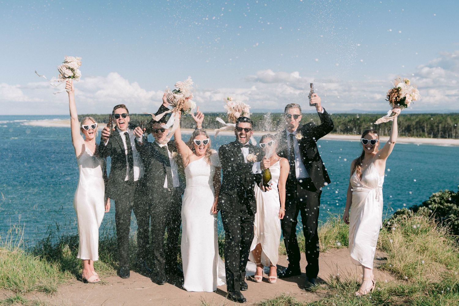Wedding party celebrates joyfully on coastal cliff overlooking blue ocean waters and beach on sunny day.