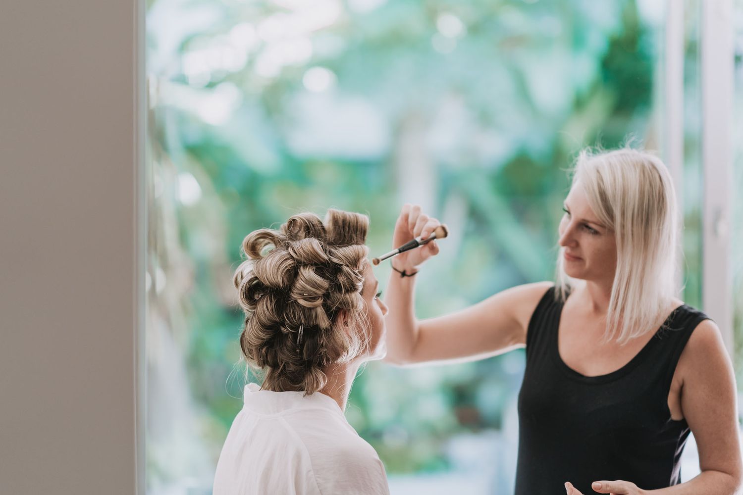 Hair stylist applying product to client's hair during bridal styling session in bright natural light setting.