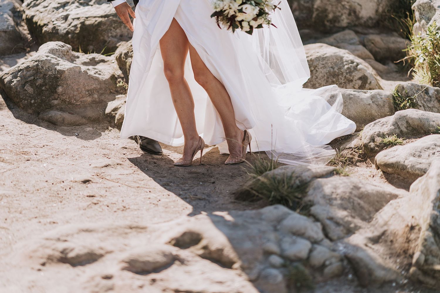 A pair of elegant white shoes sit among rocky terrain with a flowing white wedding dress in the background.
