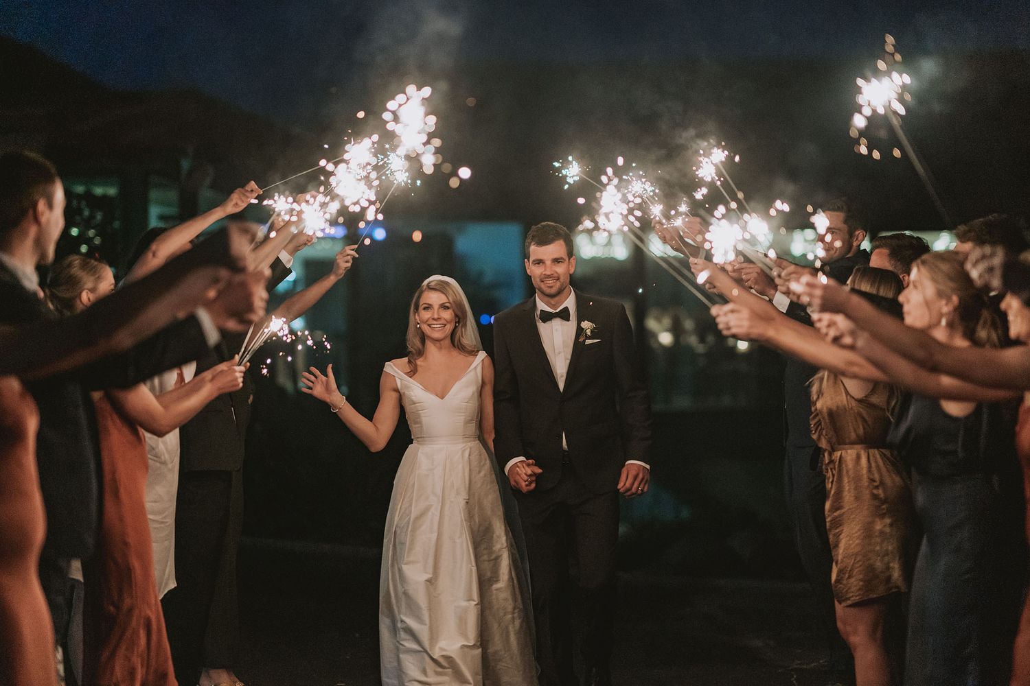 Wedding party celebrates with sparklers under the night sky during outdoor reception.