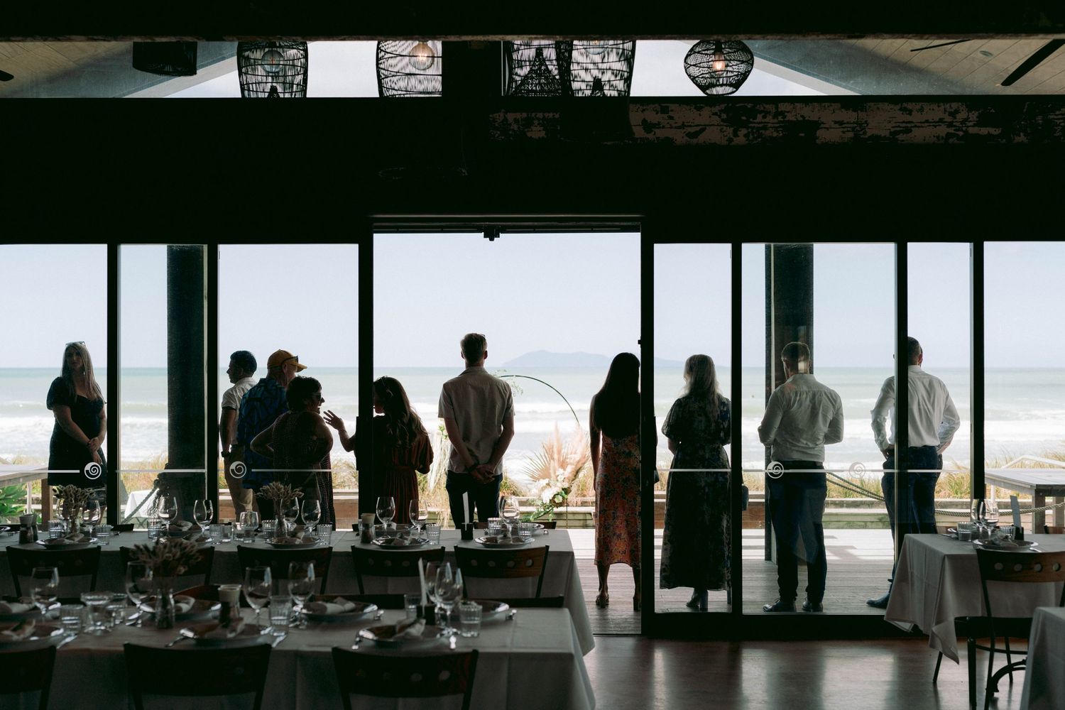 Silhouetted figures stand at large windows overlooking the ocean in a modern restaurant or bar interior.