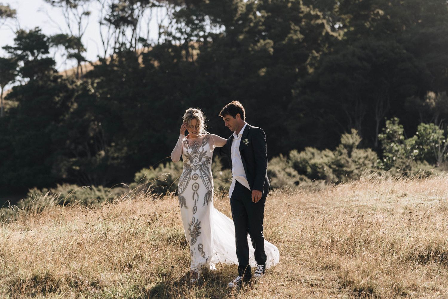 Couple in wedding attire walking together through a sun-lit field of tall golden grass with trees in background.