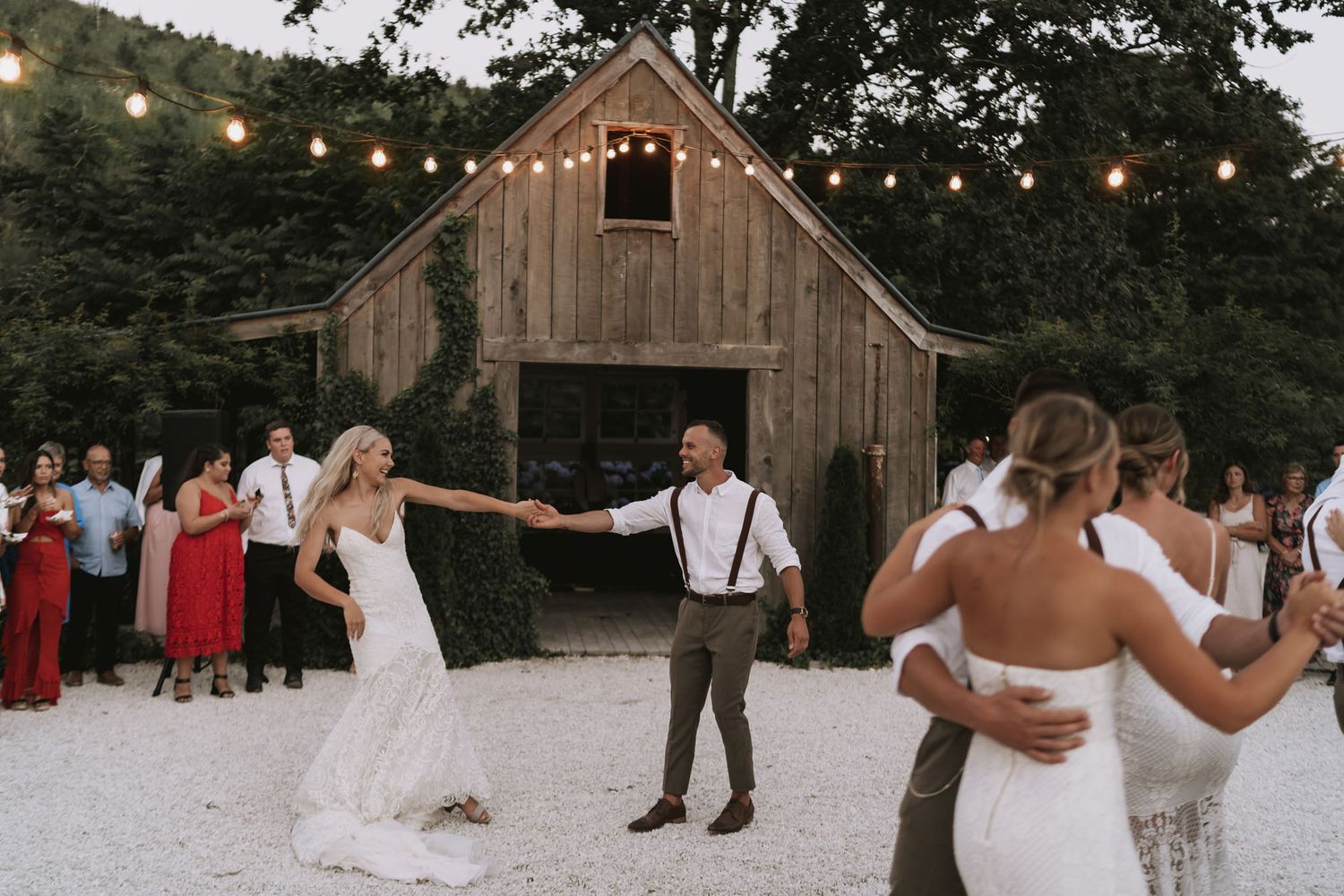 A rustic outdoor wedding reception features string lights and a wooden barn at dusk during a couple's first dance.