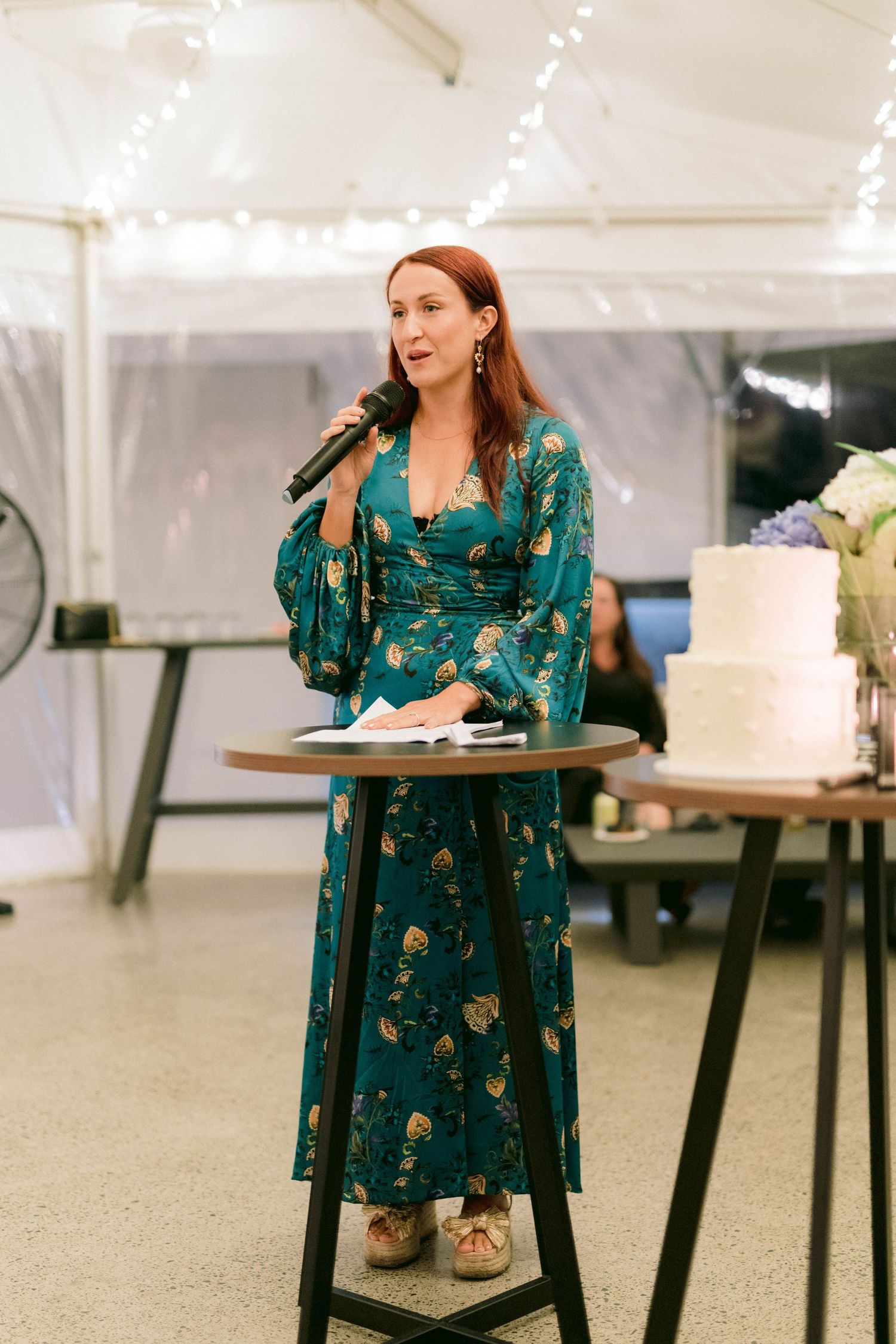 Someone in a teal floral dress speaks at a microphone stand during an indoor event with string lights and cake in the background.